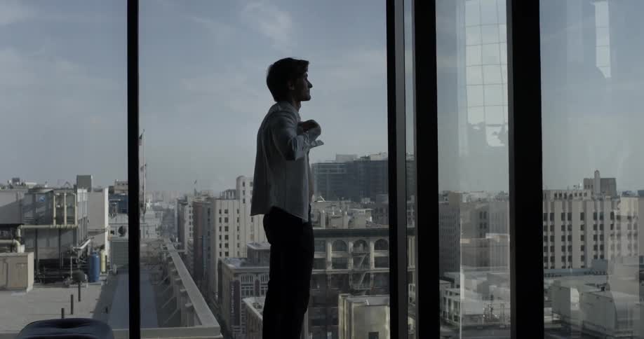 A man looks out an office window while putting on his shirt