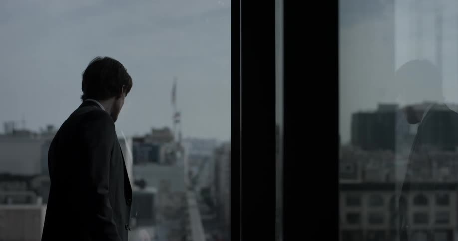 A man in a  suit looks out a window at a city
