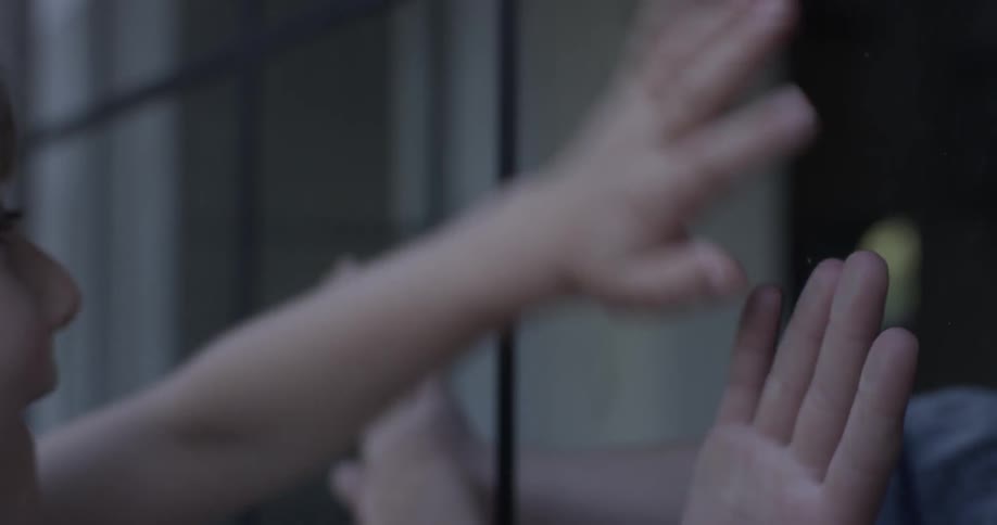 Two kids play a hand game on opposite sides of a window