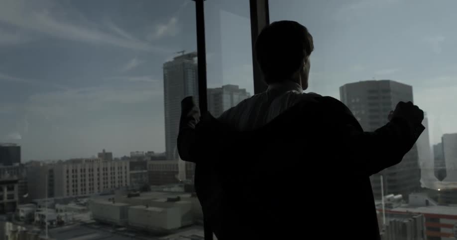 A man throws on the jacket of a suit in front of some large windows overlooking the city