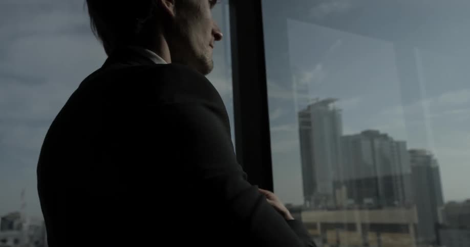 Man tosses on a black jacket in the corner of a office in front of a tall large window overlooking the city