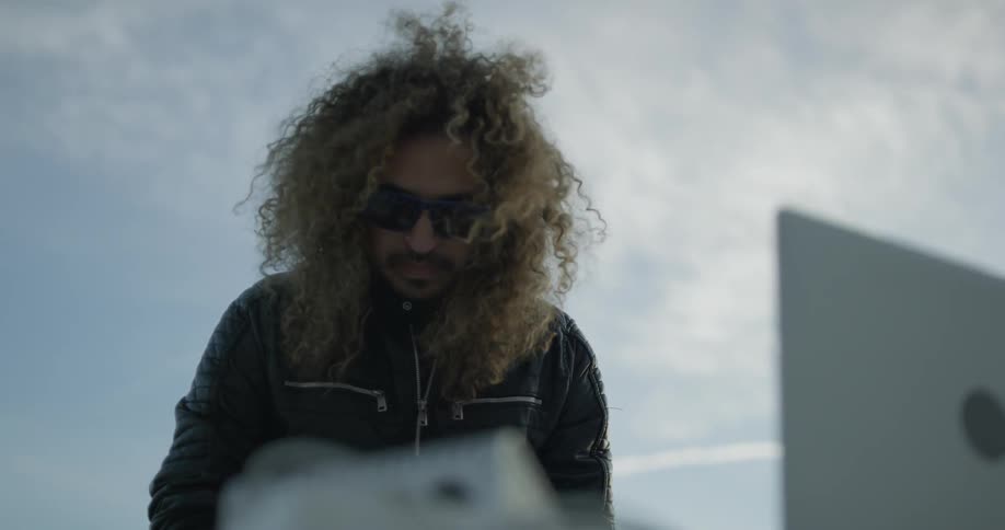 A man with sunglasses looks down at his laptop outside
