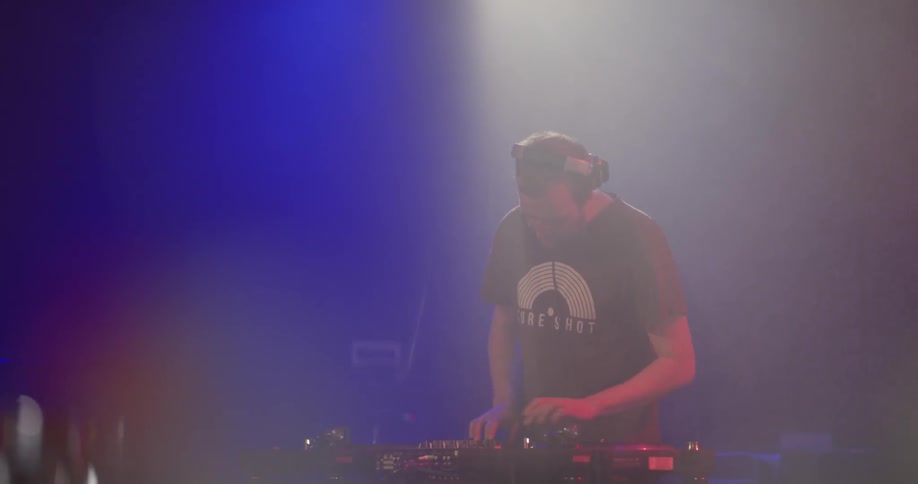 A man wearing headphones and a t shirt manages DJ equipment on a stage with red and blue lights shining