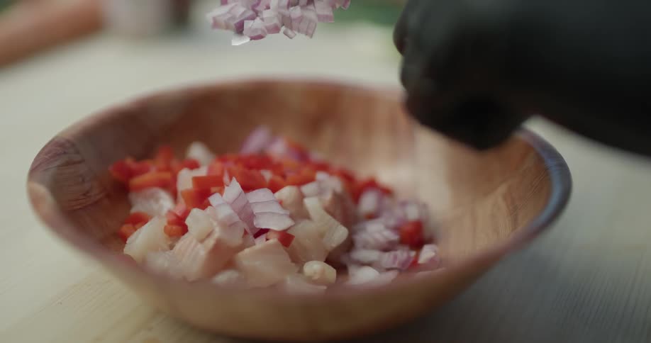 A wooden bowl with food in it has onions poured into it off of a knife help by a hand with black gloves