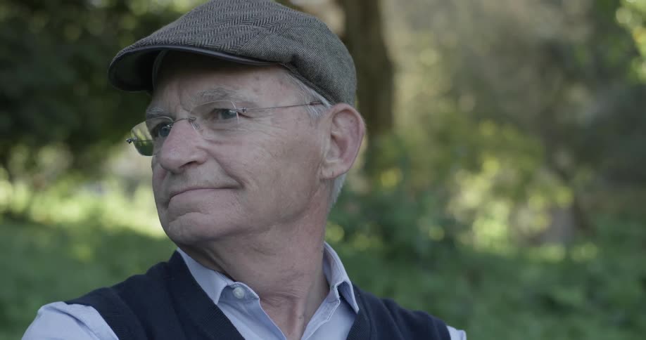 A man smiles while sitting on a park bench wearing a hat and vest and sunglasses
