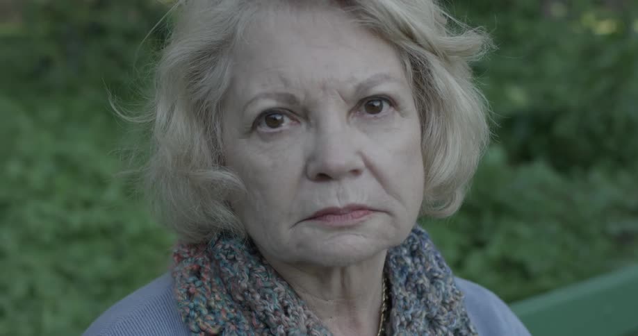 A woman sits on a bench at the park and stares at the camera with a blank stare on her face