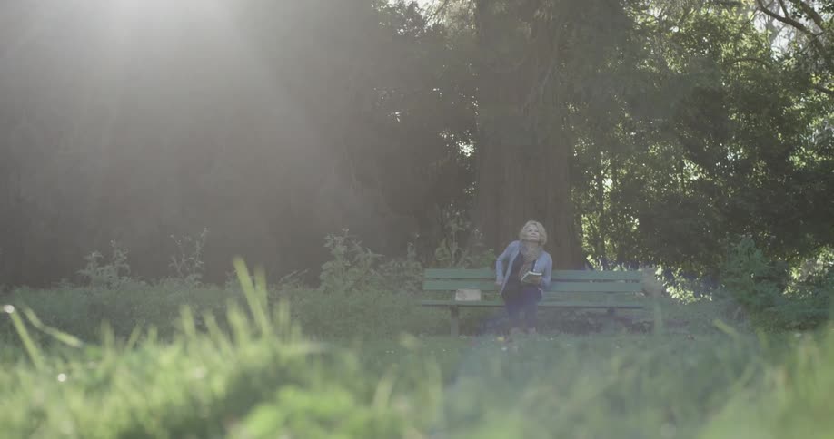 A woman walks towards a bench in the park and proceeds to sit there and put her purse down to read a book