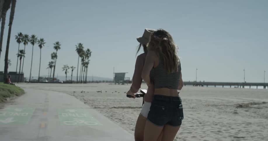Two women ride a scooter together by the beach