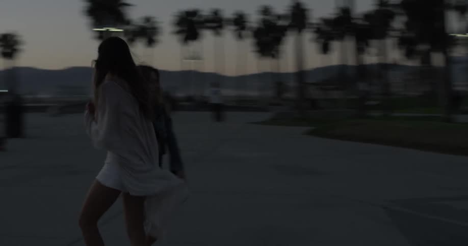 Two girls dance happily on the sidewalk after sunset near the beach with palm trees shorts and sandy shores