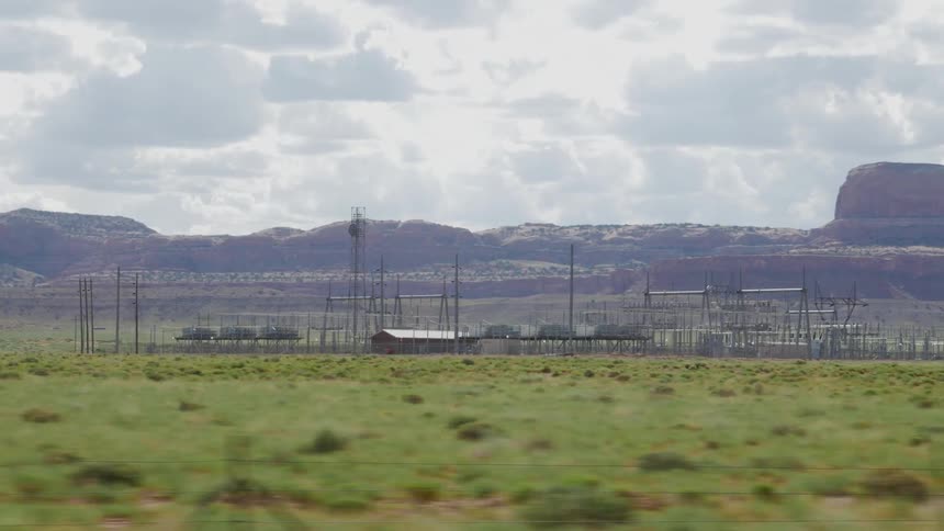 Looking out a car window at a small power station in a field, with buttes and clouds in the background