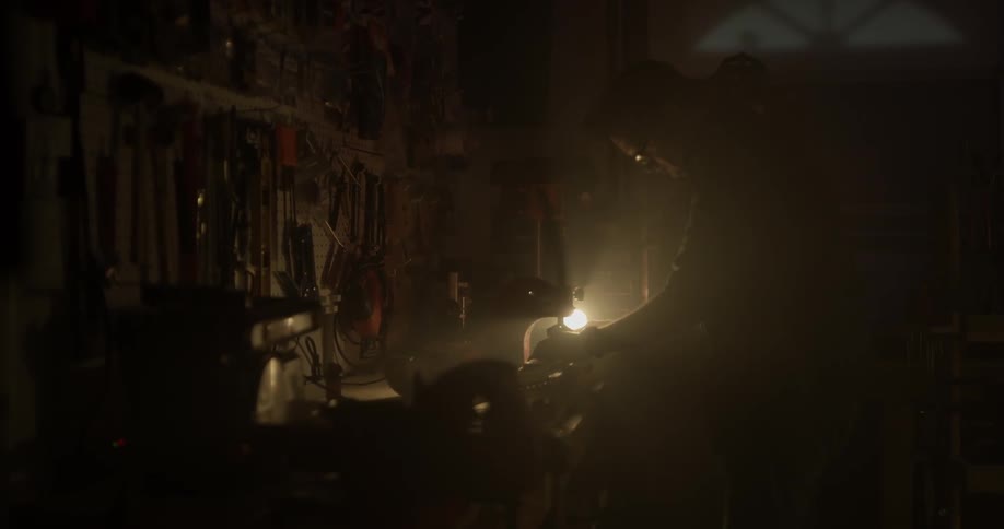 A man walks through a dark workshop to a light and then turns it on illuminating the space