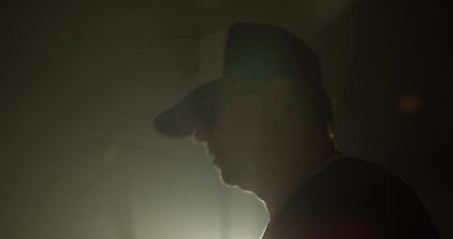 Man in a hat with sunglasses and a black shirt bangs on on a drum set in a dim lit room with a spot light