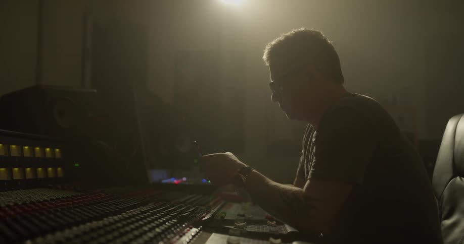 A man sitting in front of a music studio panel does something on his phone