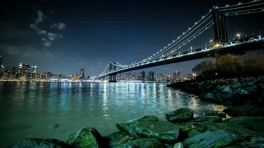Time lapses as clouds pass over bridge that spans water toward city at night