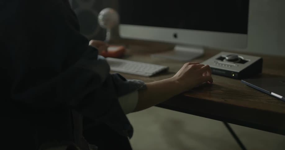 A woman is sitting at her wooden desk moving the mouse around in front of a mac monitor with white yeti snowball microphone and video studio equipment and large speakers