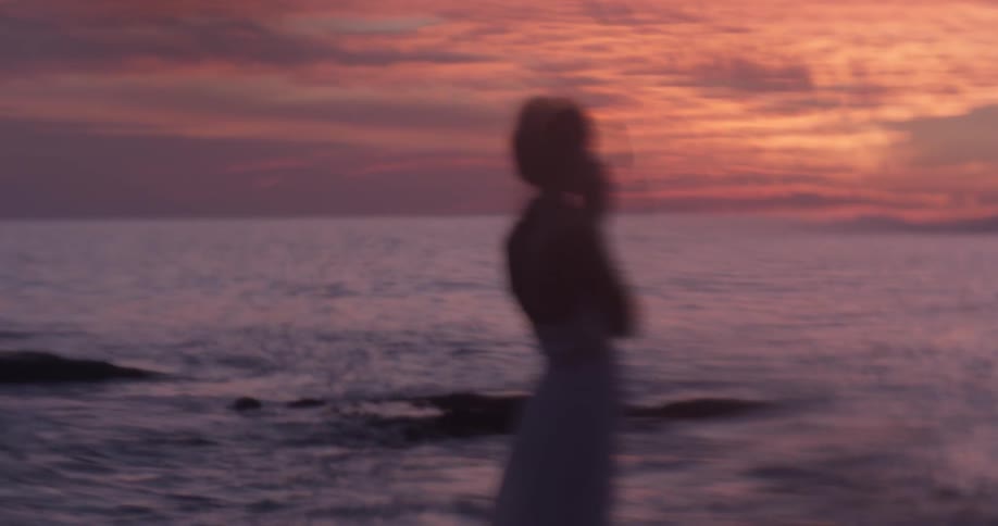 A woman stands looking out at a sunset over the ocean
