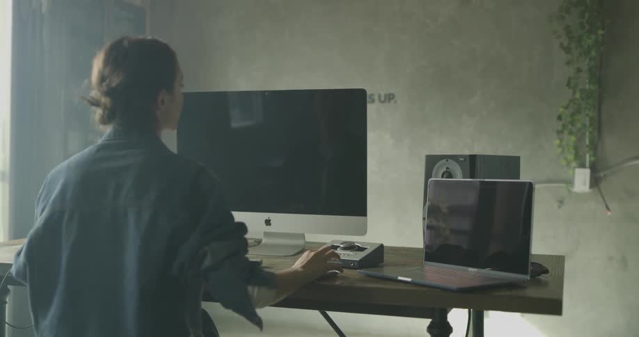 A woman sits at a computer desk clicking the mouse and typing on the keyboard on a bright sunny day indoors