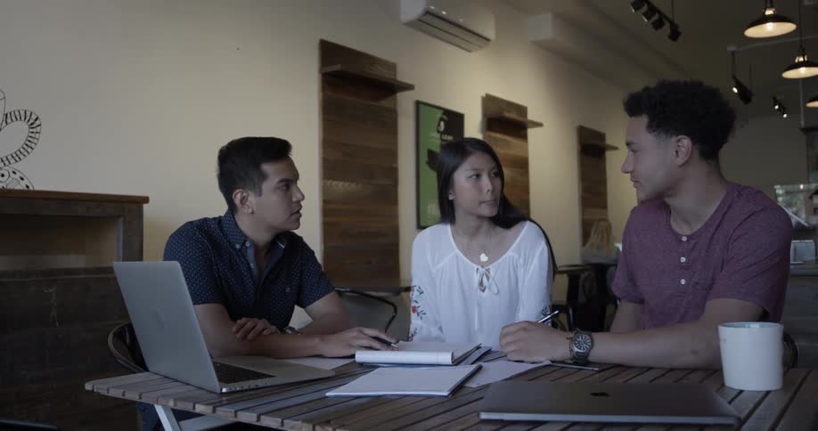 Two men and a woman discuss something over a laptop and notebooks on a table