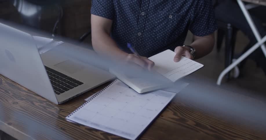 A man types on a silver laptop then writes in a notebook