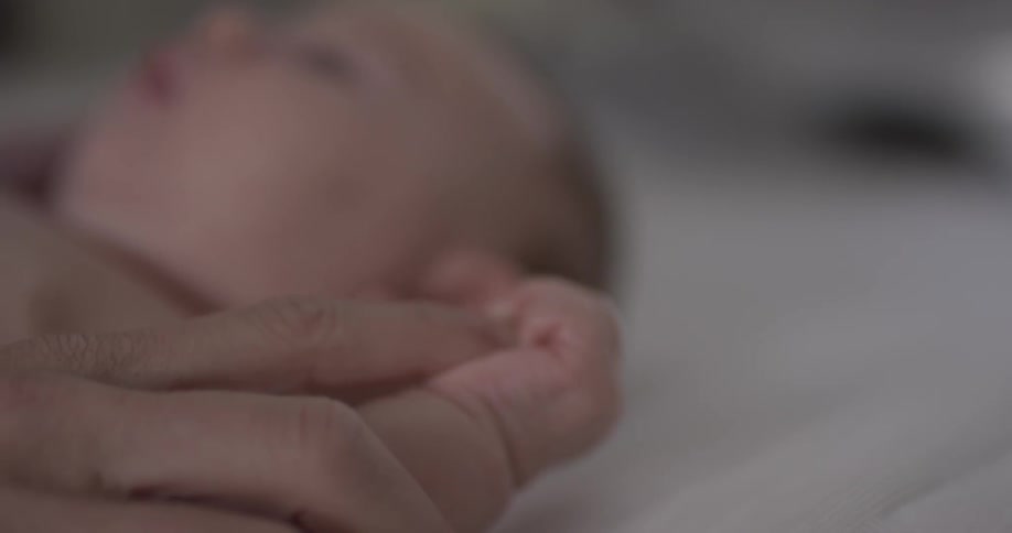 Baby is caressed on the wrist and hands by a large adult hand while infant lays down on a bed sheet