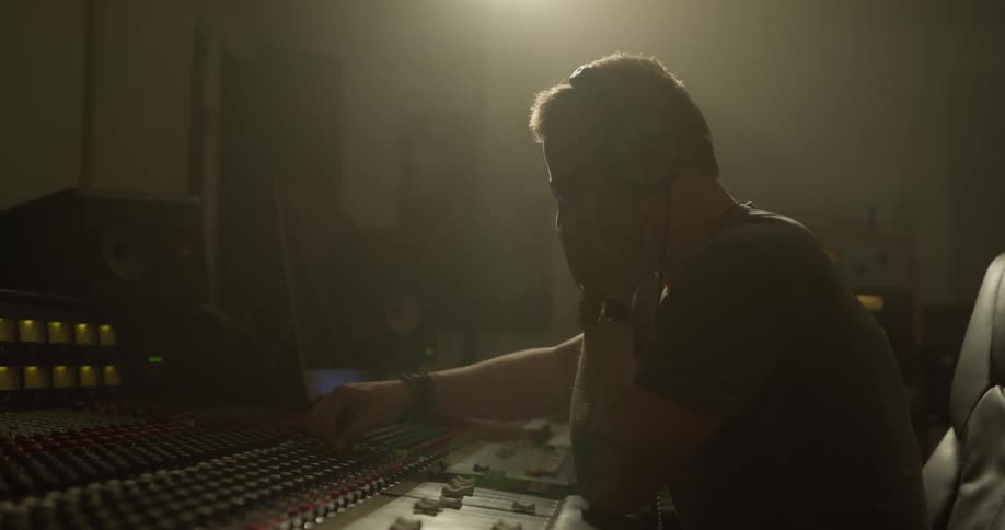 A man puts on headphones as he sits adjusting a mixing board