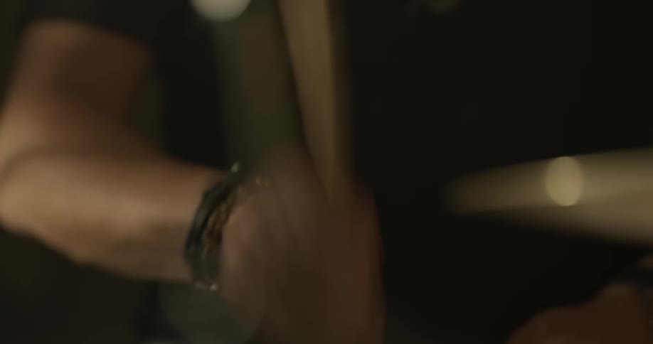 Man drums on a drum set wearing a wrist band and a black t shirt in a dim lit room
