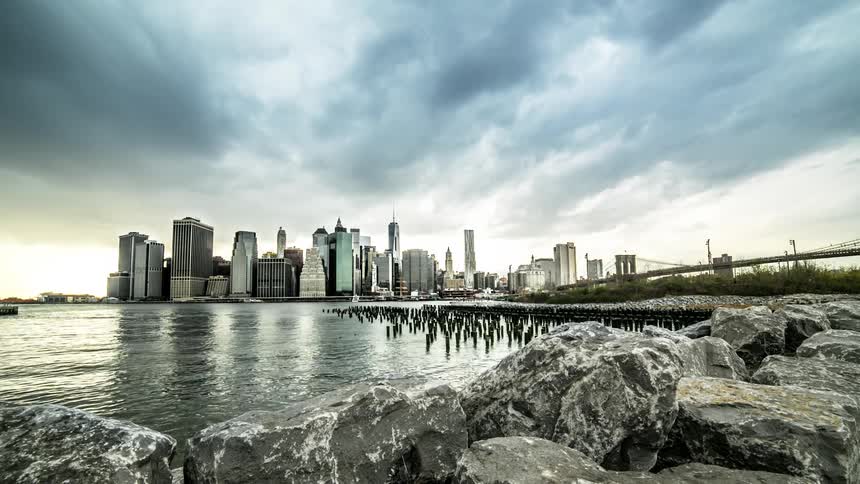 Rocks line water as clouds pass over city in the distance