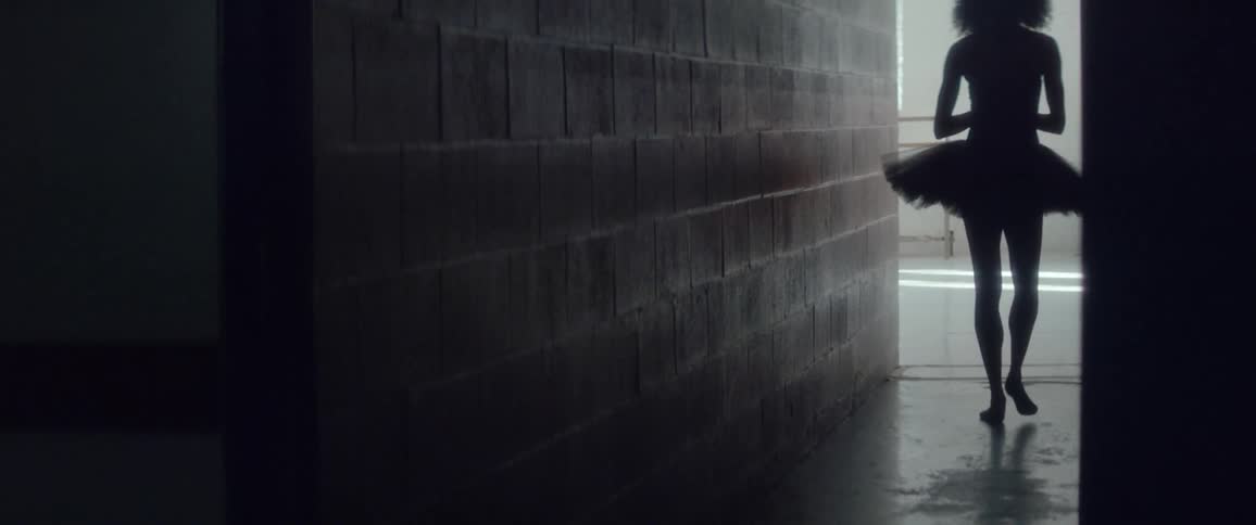 A ballerina walks down a dark hallway towards a sun lit dance studio