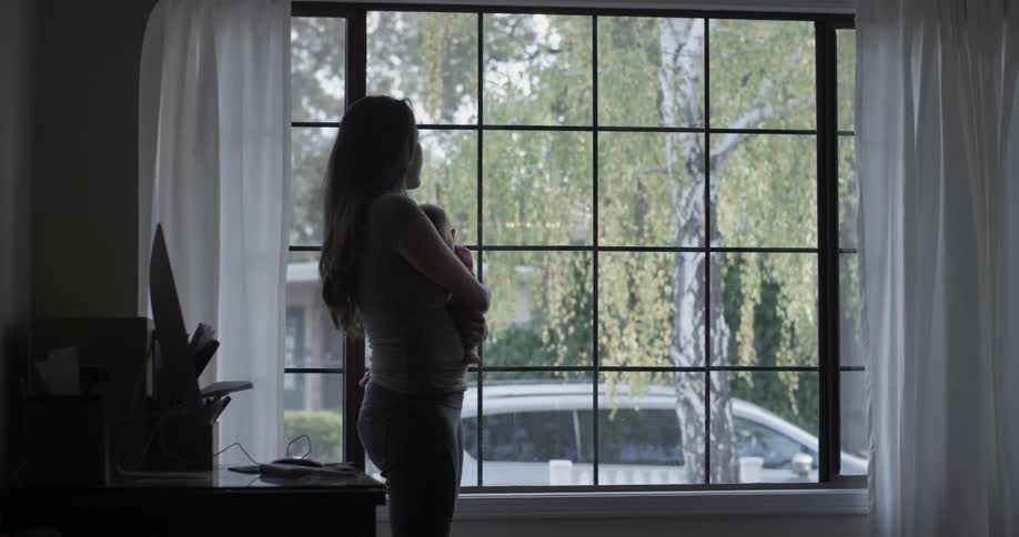 Woman holding baby stands in front of window in home