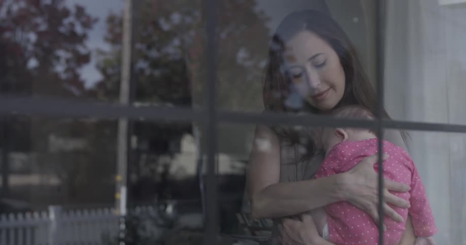 Woman cuddles baby close and stands near window during the day
