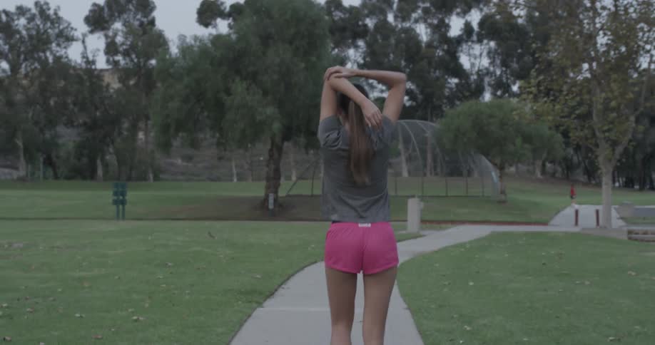 Woman in pink shorts stretching and walking down a sidewalk in a cloudy park