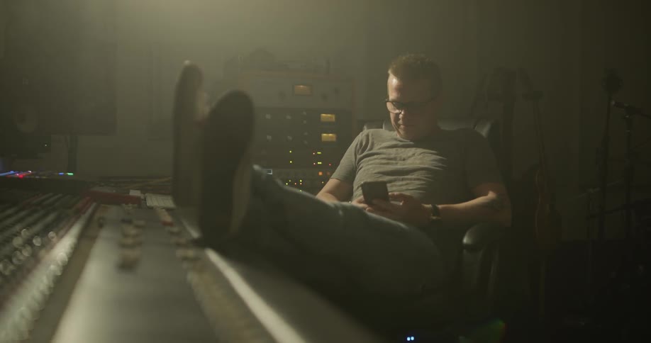 A man is sitting in a dim lit room kicking his feet up on the studio mixing board looking down at the cellphone he is holding