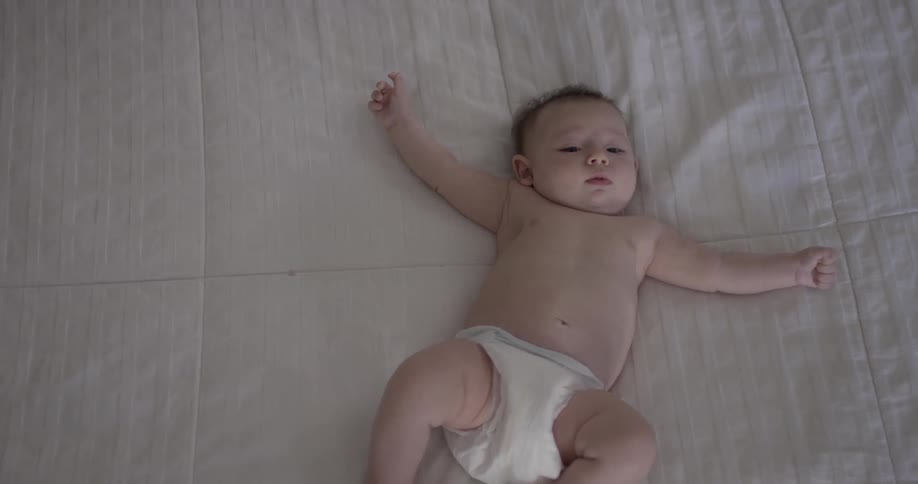 A baby lays on it's back in the middle of a white bedspread