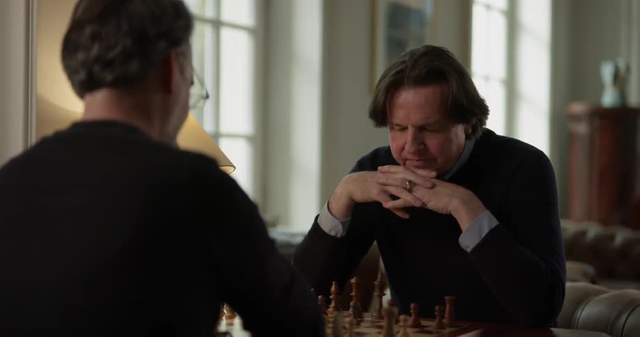 Two men one with glasses both wearing sweaters sit over a chess board made of wood inside a sophisticated apartment or home and make moves