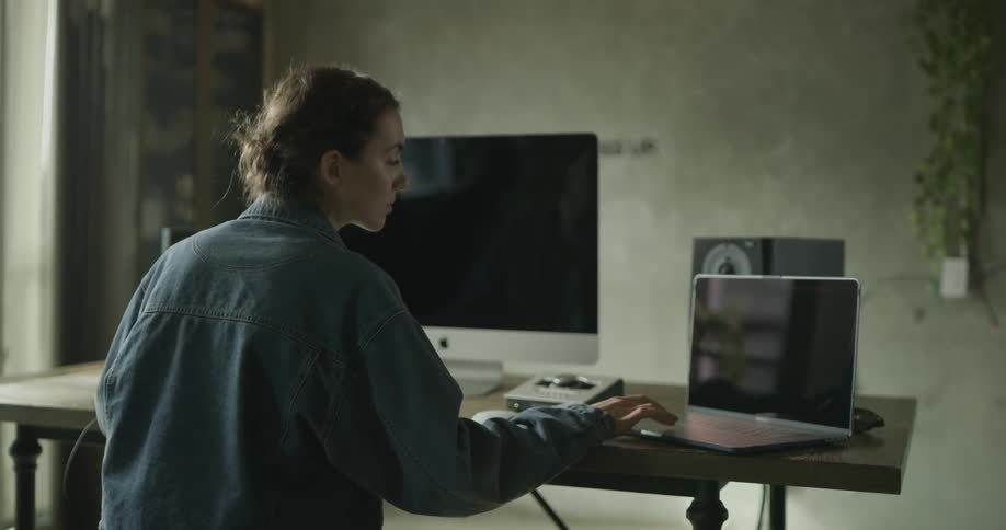 A woman sits at the wooden computer desk with a laptop and a mac computer looking at multiple screens