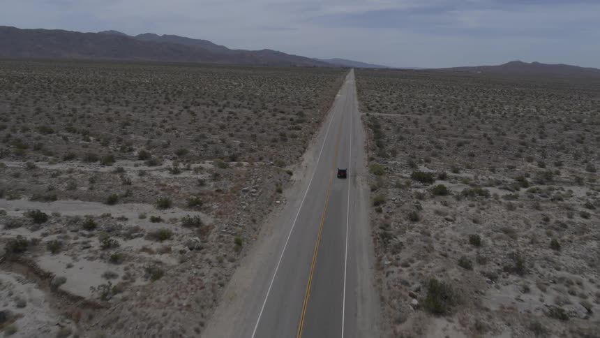 Car drives along straight road in desert with hills in the distance