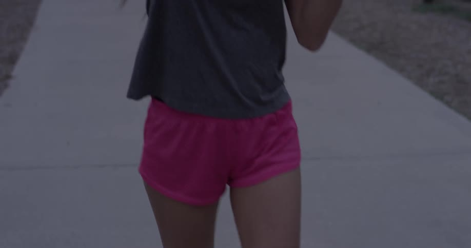 Young woman in pink shorts and a grey shirt stretching while walking on a sidewalk
