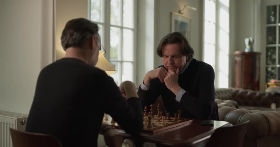 Two men play chess in living room under table lamp