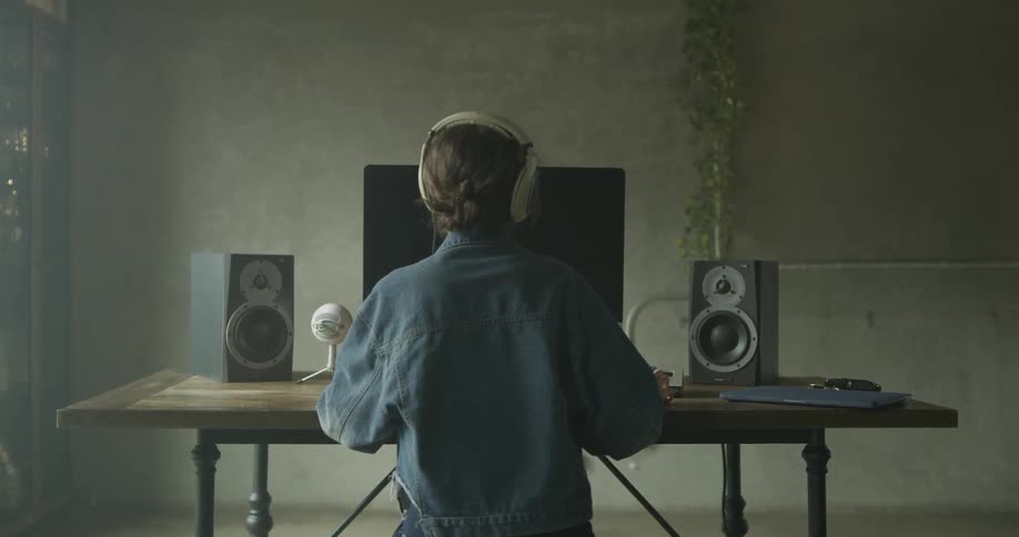 A woman wearing jeans and a jean jacket puts on white headphones in front of a desk with a snowball microphone and large speakers and mac monitor in a bright sunny day lit room