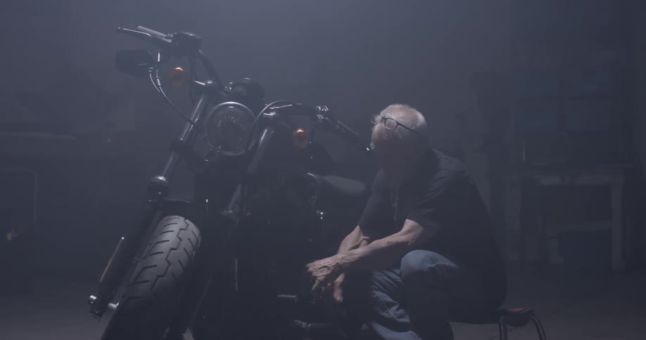 Man is in the workshop repairing a motorcycle perhaps in the garage in a dim lit room
