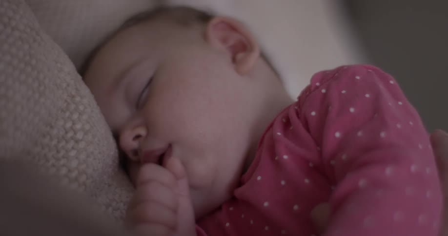 A baby nearly sucks her thumb while lying comfortably in a blanket wearing a pink shirt with white polka dots and smiles
