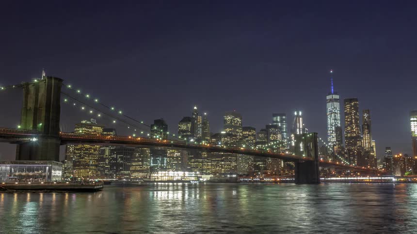 Boats float down the river next to a well lit city at night