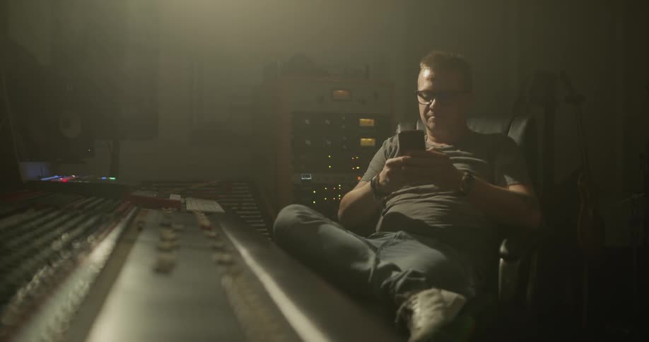 Sitting in front of a mixing board on a computer chair looking at phone wearing glasses in a dim lit room