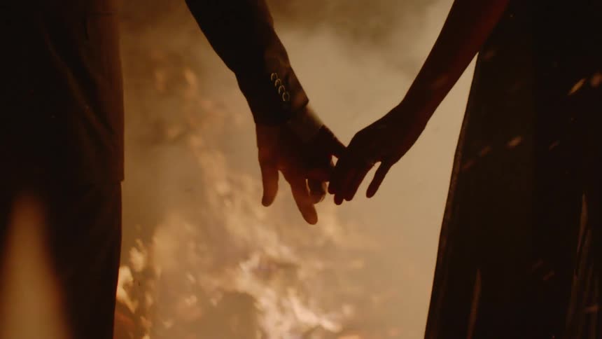 A man and woman hold hands as they watch a large fire burn at night