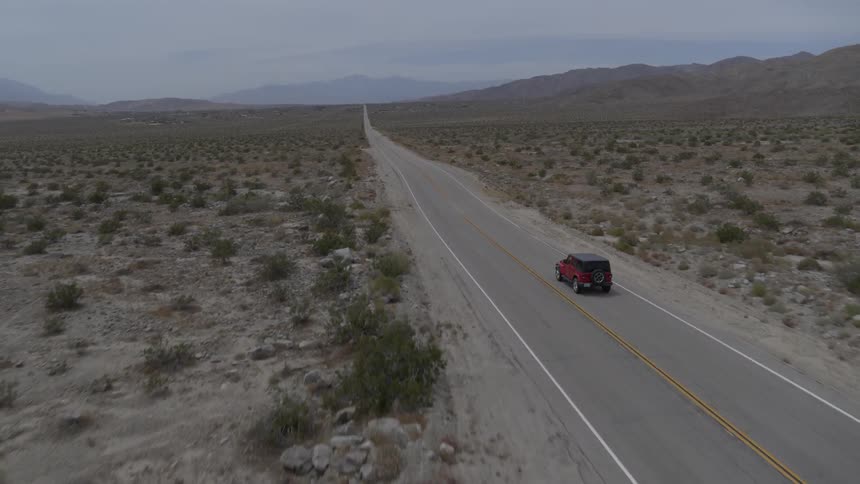 A red jeep is driving down a barren highway out in the middle of the desert with minimal vegetation on a bright cloudy but sunny day