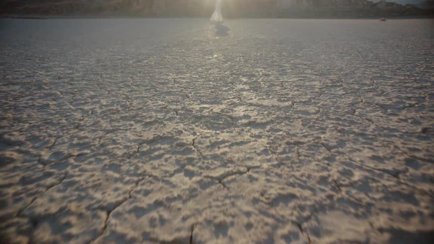 The camera rushes towards the female standing alone in the desert filled with dry cracked soil and she has a white dress on as the sun is setting