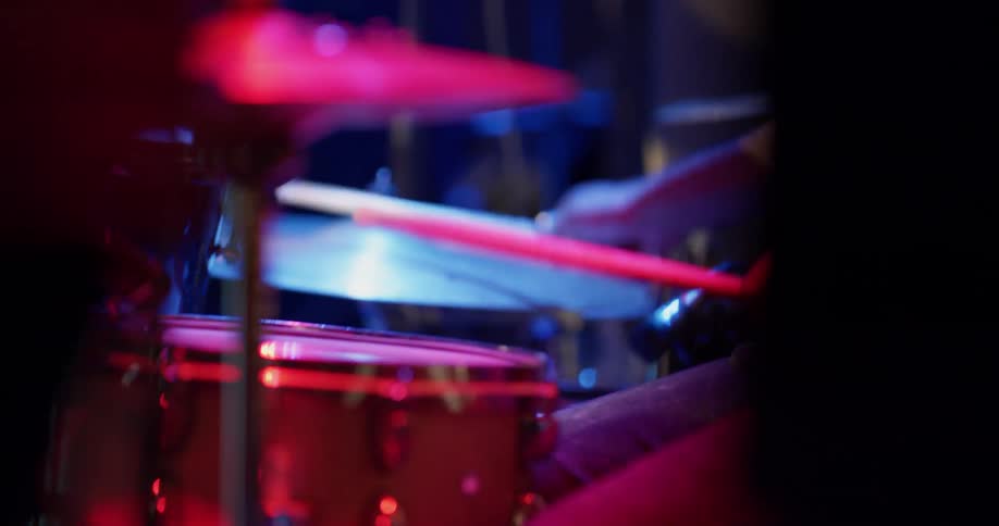 Red and blue lights shine on a drum kit on a stage as a man bangs on them with a drumstick