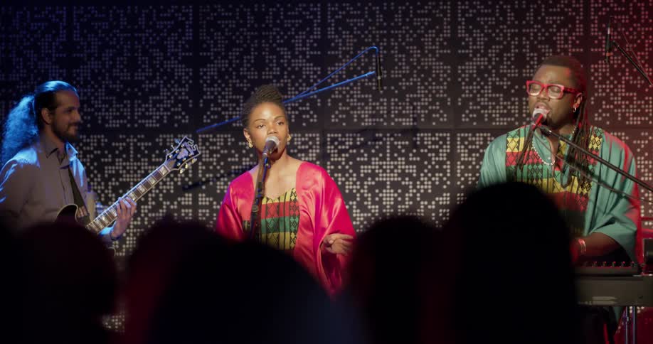 A bunch of African musicians sing and play guitar and piano on stage at a concert with red and blue lights and a spotlight