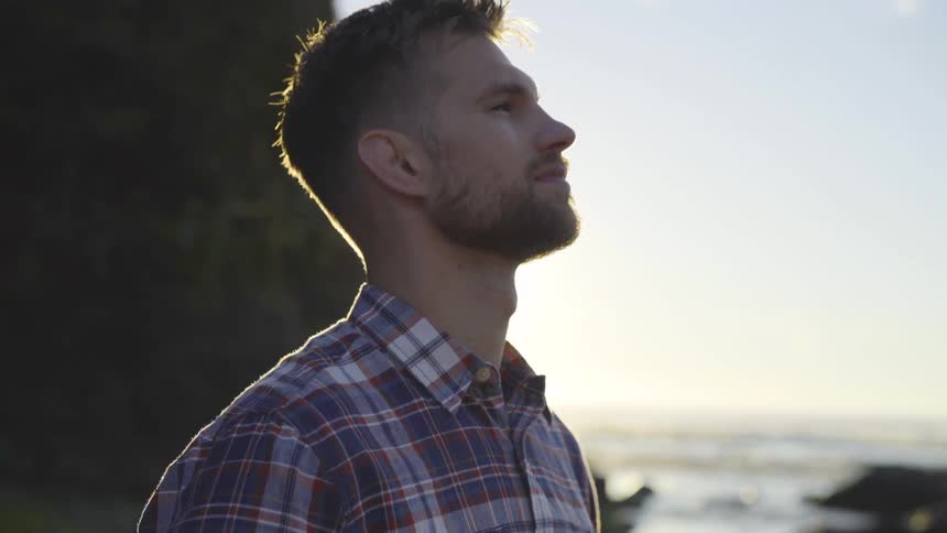 A man stands on the beach with the ocean in the background and rocks with the sun eclipsing behind him and blue skies on a sunny day