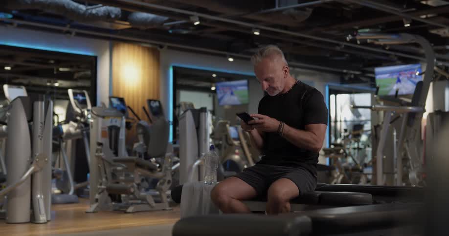 Man in gym sits on bench and checks his phone